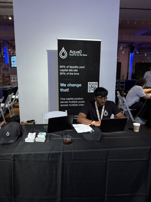The image shows a man sitting at a table with two laptops open in front of him. He appears to be working on one of the computers while wearing a black shirt. The other laptop is placed next to his, suggesting that he might be multitasking or comparing information between the devices. In addition to the laptops, there are several books and papers scattered around the table, indicating that the man could be researching or studying something related to cryptocurrency. A sign on the wall behind him reads "Aqua0", which is likely a reference to the company they are promoting at their booth.