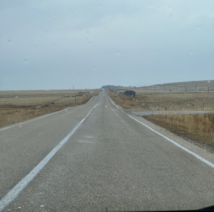 A view from a car windshield of a long, straight highway stretching through a flat, barren landscape under an overcast sky.
