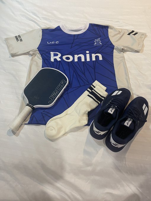 A flat lay of sports gear on a white sheet: a blue 'Ronin' jersey (UCC logo), a JOOLA pickleball paddle, white socks, and navy blue athletic shoes.
