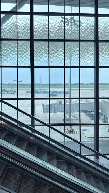 A view through a large grid-patterned window inside an airport terminal, showing an airplane taking off, a jet bridge, and an empty staircase in the foreground.