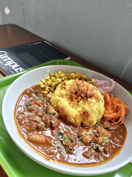 A bowl of yellow rice topped with crispy shallots sits alongside a rich brown curry with meat, accompanied by diced vegetables and shredded carrots on a green tray.