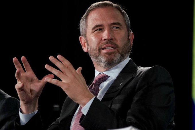 A middle-aged man with a beard, wearing a dark suit and tie, gesturing with his hands while speaking against a dark background.