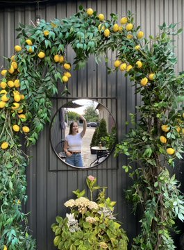 A decorative garden scene featuring a round mirror framed by a leafy arch with yellow lemons, mounted on a dark wall, with potted plants and flowers in the foreground.