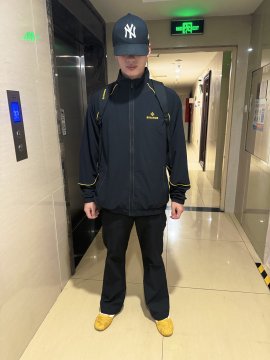 A person wearing a navy NY Yankees jacket and cap stands in a hallway, paired with black pants and yellow shoes.