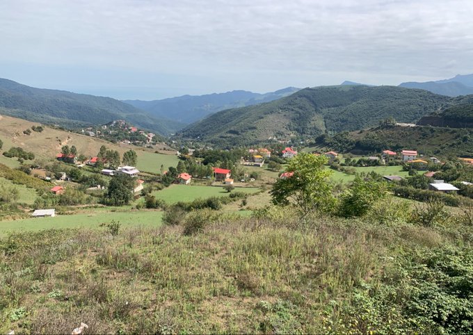 A scenic view of a green, mountainous valley with scattered houses and lush vegetation under a hazy, cloudy sky.