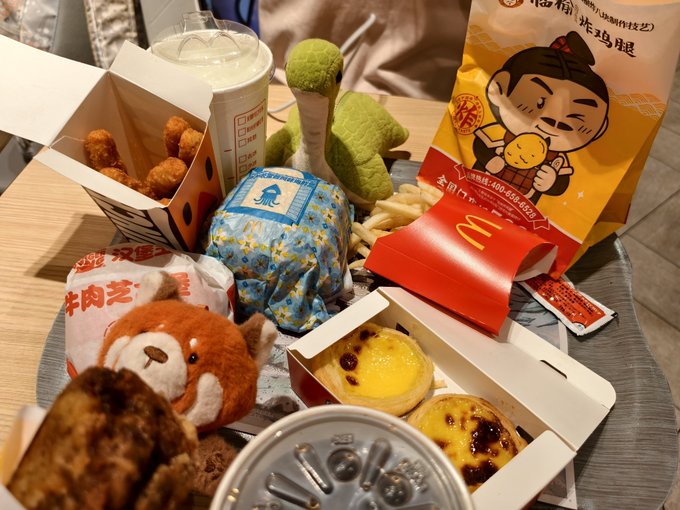 A McDonald's feast spreads across the table, featuring fries, a cheeseburger, egg tarts, and a McNugget bag with Chinese text "炸鸡腿" (fried chicken leg), accompanied by cute plush toys. The post's poetic couplet "东市买薯条西市买蛋挞" (fries from the east market, egg tarts from the west) humorously describes the variety of McDonald's items pictured, celebrating a multi-course fast food haul.