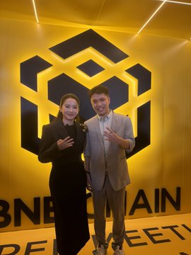 Two people posing in front of a yellow wall featuring the BNB Chain logo, with text reading 'BNB CHAIN' and 'MEET' visible behind them.
