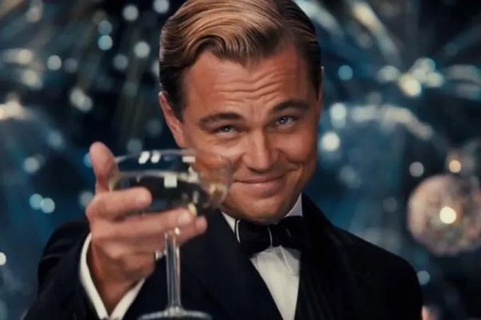 A dapper man in a tuxedo smiles warmly while raising a champagne glass in a celebratory toast, against a backdrop of glittering lights and disco balls. The image pairs with the post's cheerful "happy Saturday!" message, capturing a spirit of festive enjoyment reminiscent of a glamorous Gatsby-era party.