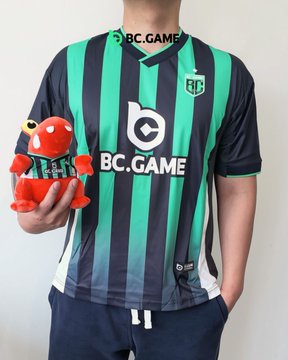A person wears a green and black vertically striped BC.GAME soccer jersey featuring a crest with 'AG' initials, holding a small red robot plush toy.