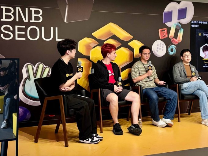 Four men sit on stage chairs with microphones during a panel discussion at a 'BNB SEOUL' event, with colorful honeycomb-themed and gaming icon decorations behind them.