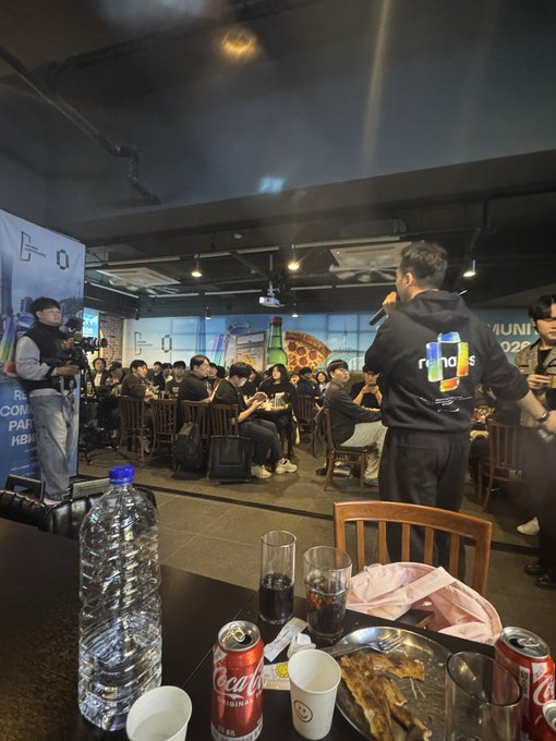An indoor event space with a crowd seated, featuring a person in a 'namUs' hoodie speaking, with food, drinks (including water bottle and Coca-Cola), and a cameraman filming in the foreground.