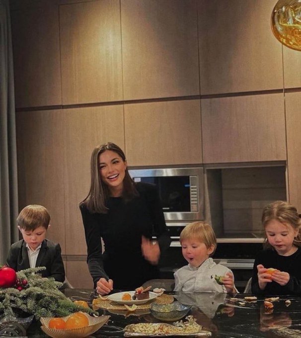 A photo of a smiling woman with three young children in a modern kitchen, gathered around a counter with various snacks and Christmas decorations.