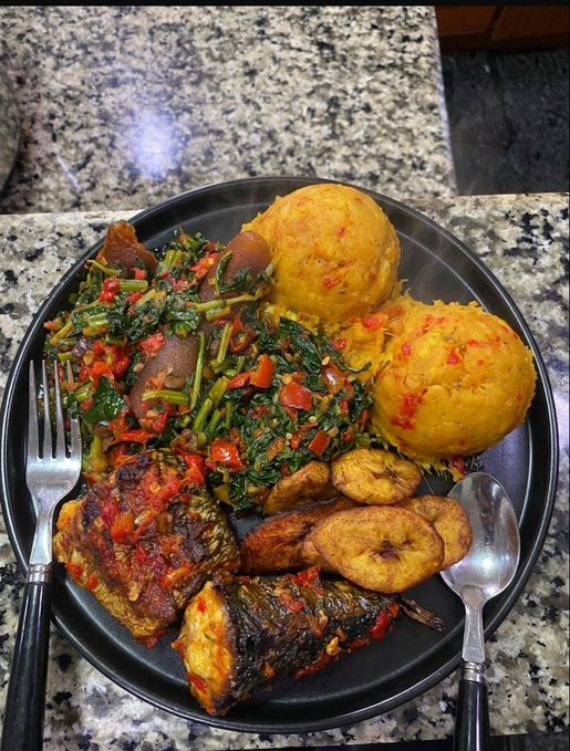 A plate of homemade West African/Afro-Caribbean cuisine featuring yellow garri or eba-style mounds, sautéed greens with peppers, fried plantain slices, and grilled fish with tomato-pepper sauce, paired with a fork and spoon — a hearty homemade dinner matching the caption's invitation to share what others are eating.