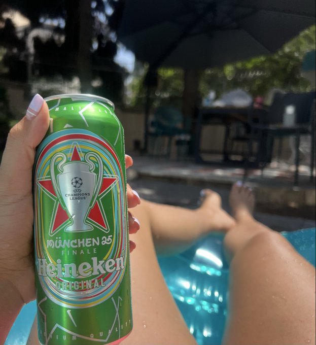 A hand holds a green Heineken beer can commemorating the UEFA Champions League München 25 Finale, with legs visible by a poolside in the background.
