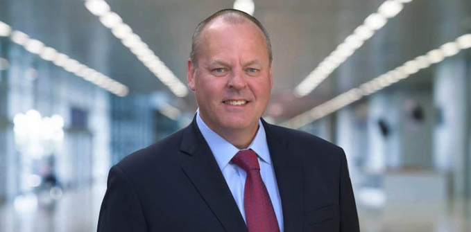 Professional headshot of a smiling middle-aged man in a dark suit with a blue shirt and patterned red tie, photographed in a modern building interior.