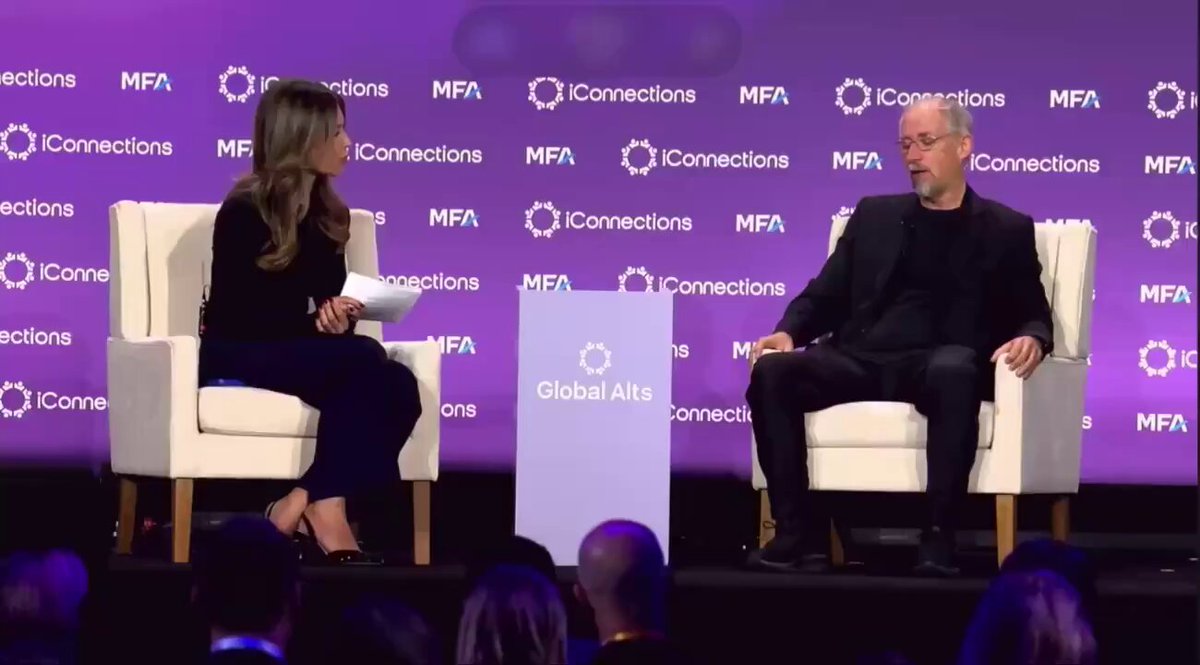 The image shows a man and woman sitting across from each other at a conference stage, engaged in conversation. They are positioned behind a podium that displays the text "Global Connections". The backdrop features purple and white text, possibly indicating an event or gathering related to global connections. A crowd of people can be seen in the background, suggesting this is a public event with attendees listening to the speakers.