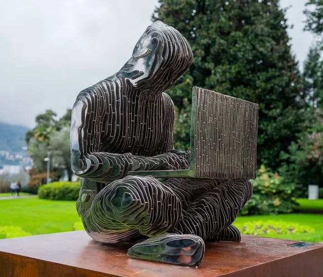 A layered, metallic sculpture of a hooded figure sitting cross-legged while holding a laptop sits on a pedestal in a park setting.