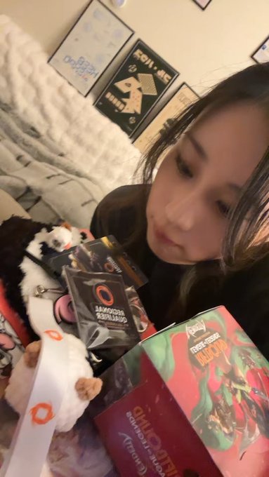 A young woman lies on a bed showing off Overwatch 2 merchandise including a Genji collector box, branded snack bags, and a small plush toy. Framed artwork is visible on the wall behind her.