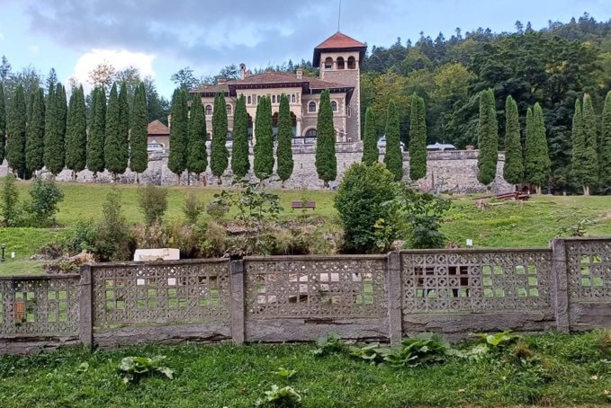 A large stone building with a tower and red roof sits on a hill, surrounded by tall cypress trees and accessed via a decorative concrete fence.