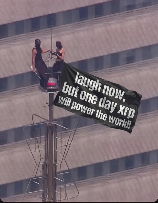 Two people on a tall pole/structure holding a green banner that reads 'laugh now, but one day xrp will power the world!' with a building visible in the background.