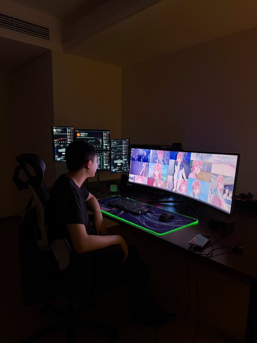 A person sits in a dark room at a desk equipped with multiple monitors, a large curved screen showing anime, and a glowing RGB mousepad.