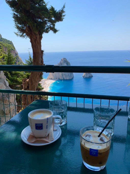 A scenic ocean view from a balcony table features a cappuccino in a 'Living Larder' cup, an iced coffee, and water glasses, overlooking turquoise waters and rocky cliffs.