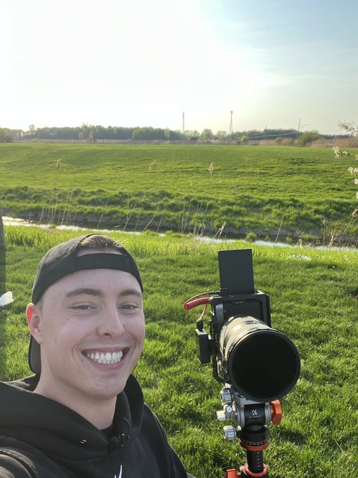 A smiling young man wearing a backward cap poses for a selfie next to a professional camera mounted on a tripod, set in a green grassy field.