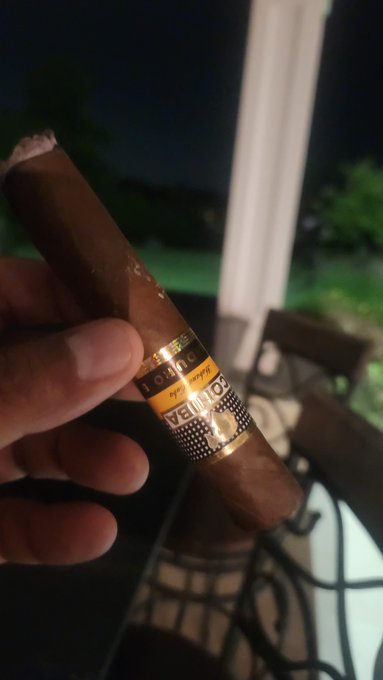 A first-person perspective photo of a hand holding a lit 'COHIBA ROYALE' cigar outdoors at night, with a patio table visible in the blurred background.