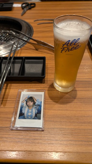 A glass of "All Free" beer sits on a wooden restaurant table next to a built-in grill, with a polaroid photo of a young woman in a blue patterned sweater displayed nearby.