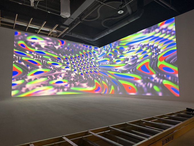 An immersive art installation displays colorful, undulating geometric patterns projected across two large walls in a corner room.