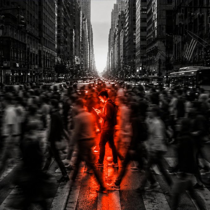 A young man stands still, illuminated by a glowing red light from his phone, amidst a blurred crowd of rushing pedestrians on a city street, symbolizing how refusing to follow the ordinary pace of the world makes him stand out. The image's striking contrast between stillness and motion captures the post's message of standing apart from the crowd.