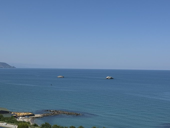 A serene morning view of a calm blue ocean with small rocky islets in the distance and a coastal area with beach umbrellas below, perfectly capturing the peaceful vibes of a "Gm Everyone" greeting on a beautiful day.