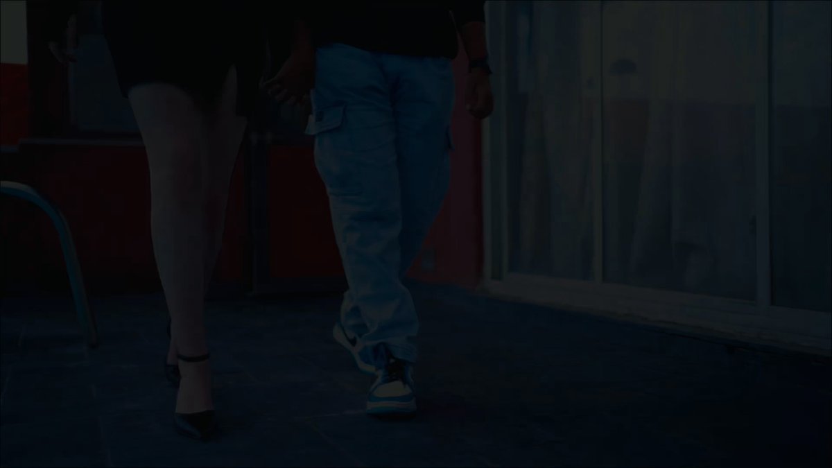 In the image, there are two people walking down a street at night. One person appears to be leading while the other follows closely behind. They seem to be engaged in conversation or walking towards something off-camera, with one person carrying a handbag that is visible as they walk together. The background of the photo provides context for their location but does not provide any additional details about the situation beyond what we can see in the frame.