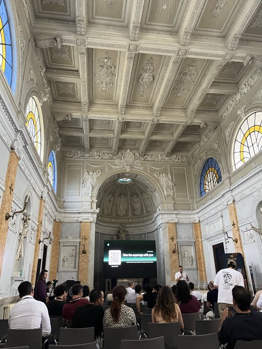 The image shows a large room with a high ceiling, featuring ornate decorations such as arches and columns. A group of people are seated in chairs facing a stage or podium at the front of the room, suggesting that they may be attending an event or gathering. The presence of multiple people indicates that this could be a social function or meeting taking place within the space.