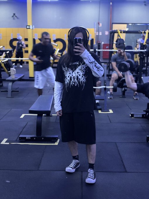 A person taking a mirror selfie in a gym, wearing headphones, sunglasses, a black graphic t-shirt over a white long-sleeve shirt, and black shorts, with other gym-goers visible in the background.