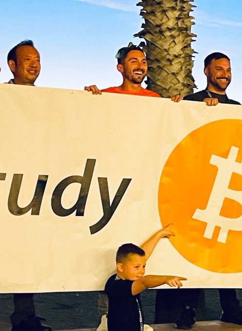 Three men stand behind a large white banner displaying part of a Bitcoin logo and the letters "tudy," with a young boy in the foreground pointing to the right.