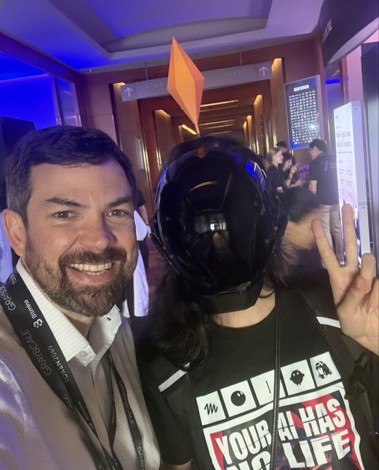 A smiling bearded man poses for a selfie next to a person wearing a large black VR/AR headset, who is making a peace sign at what appears to be a tech event.