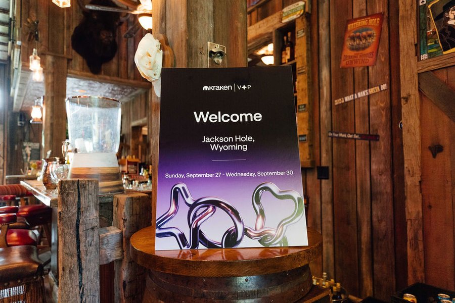 A "Kraken | V+P" welcome sign on a wooden barrel in a rustic, wood-paneled venue reads "Welcome, Jackson Hole, Wyoming, Sunday, September 27 - Wednesday, September 30," tying directly to the VIP client event hosted by Kraken in Jackson Hole that featured a ranch, summit, and private planetarium experience.