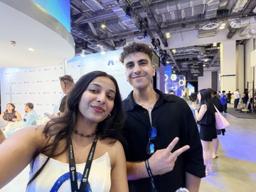 A selfie of a young woman and a young man at what appears to be a convention or expo, both wearing lanyards and making peace signs, with various booths and attendees visible in the background.