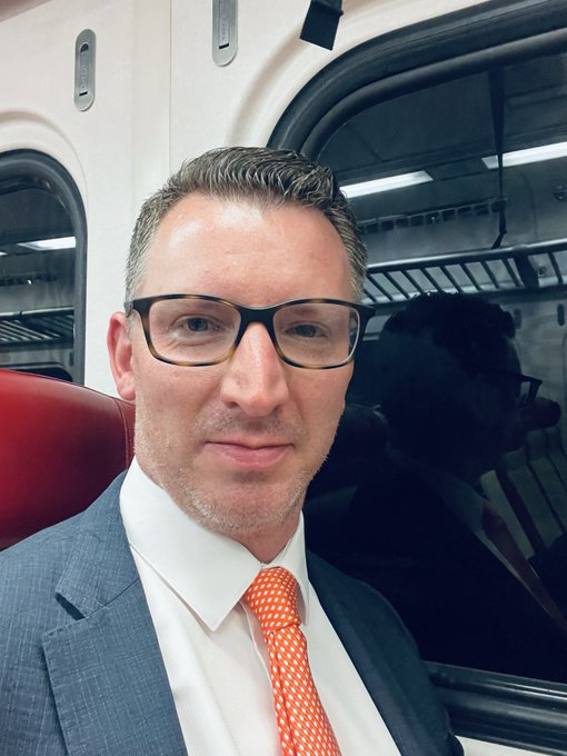 A man wearing glasses, a blue suit, and an orange tie takes a selfie inside a train car with a red seat visible.