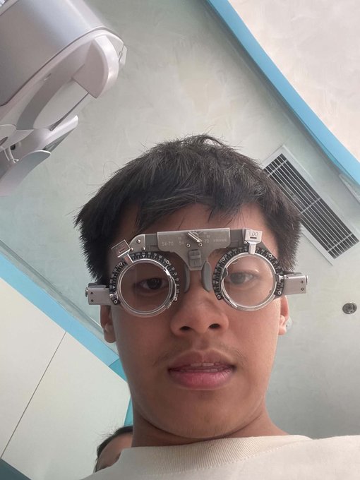 A close-up selfie of a young man wearing a trial frame with adjustable lenses, typically used for an eye exam, taken in what appears to be an optometrist's office.