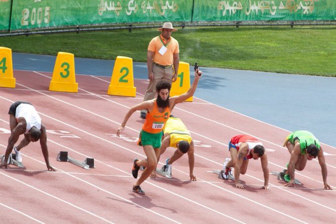 Athletes in starting positions on a red running track with lane markers, an official in an orange shirt, and Arabic text with '2005' on the green backdrop.