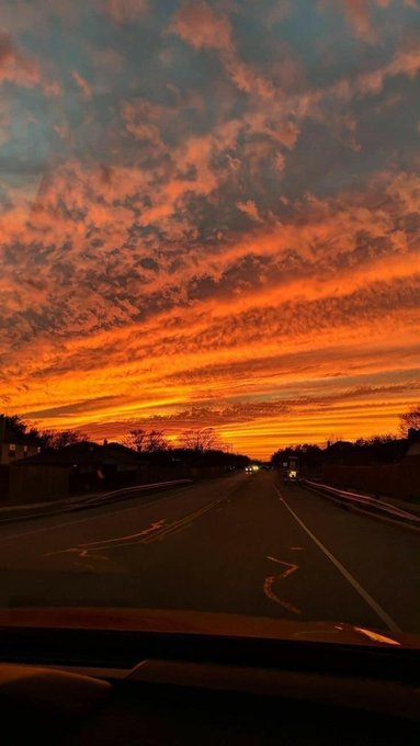 A stunning sunset view from inside a car driving down a highway, with dramatic orange and pink clouds streaking across the sky.