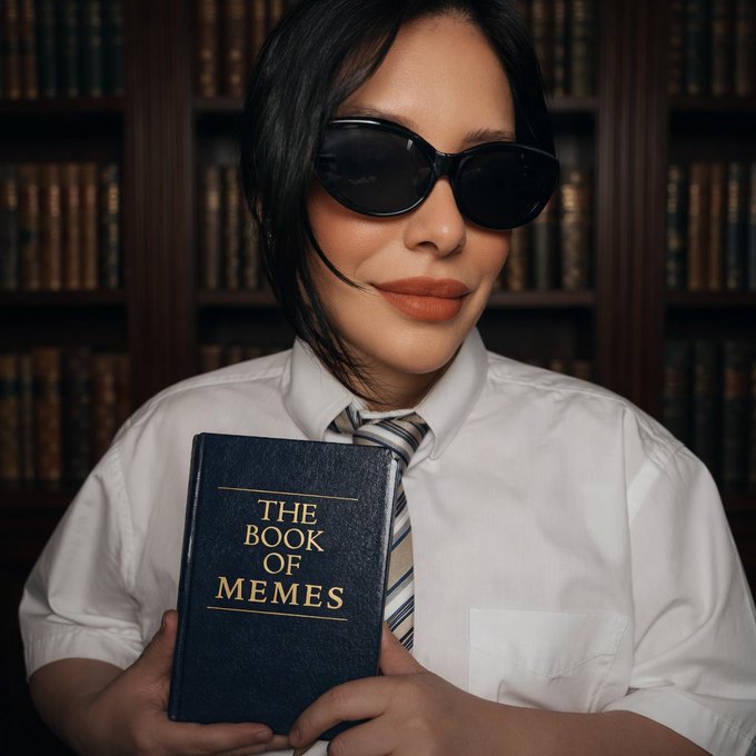 A woman in dark sunglasses and a white shirt with tie holds a book titled "THE BOOK OF MEMES" against a backdrop of bookshelves, perfectly matching the post's playful announcement that "meme season" is here.