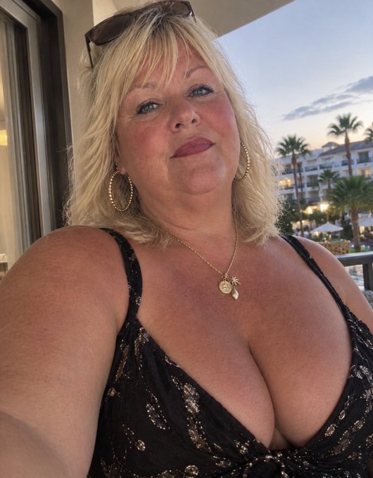 A selfie of a blonde woman wearing a black patterned top with gold necklaces and hoop earrings, posing outdoors at what appears to be a resort with palm trees in the background.