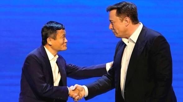 Two men in dark suits shaking hands on a stage with a blue background.