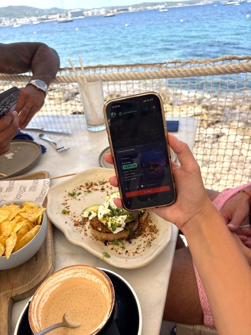 A first-person view at a seaside restaurant where someone holds a smartphone displaying a music app over a plate of food with chips and coffee.