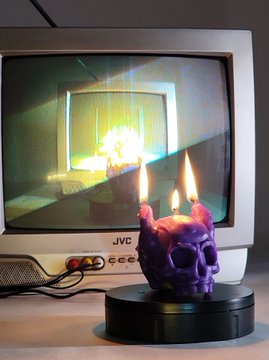 A purple melting skull-shaped candle burning in front of an old JVC CRT monitor displaying a green-tinted image.