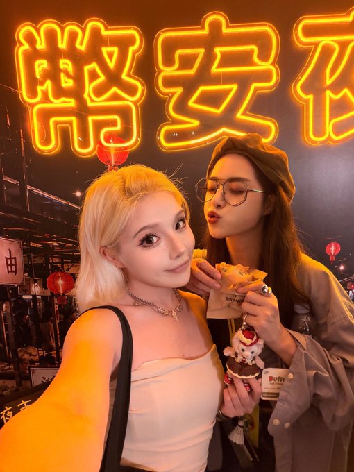 Two young women taking a selfie in front of a bright orange neon sign displaying Chinese characters, with one holding a snack.