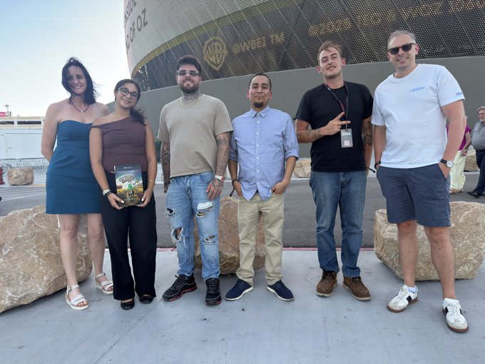 A group of six people poses for a photo in front of the Warner Bros. Studio Tour Hollywood sign, with one person holding a book.